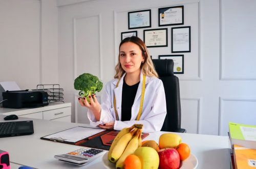 Female nutritionist in office holding broccoli, surrounded by fruits, promoting healthy lifestyle.