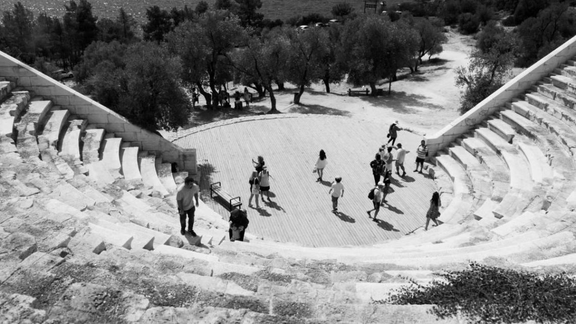 Black and white photo of people exploring an ancient Greek amphitheater by the sea.