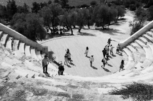 Black and white photo of people exploring an ancient Greek amphitheater by the sea.